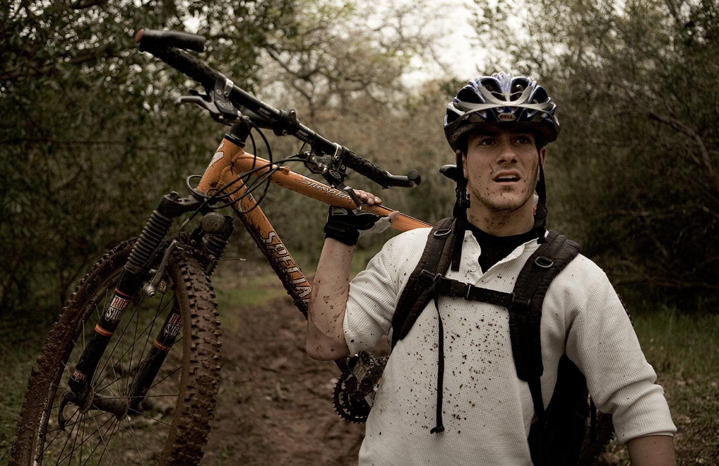 Image of Man in White Shirt with Biking Apparel Lifting Bike Behind Him Against Background of Forest