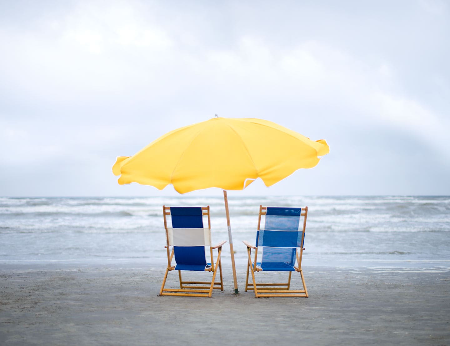 Two Blue Beach Chairs and a Yellow Sun Umbrella Facing Water Image Perspective Behind