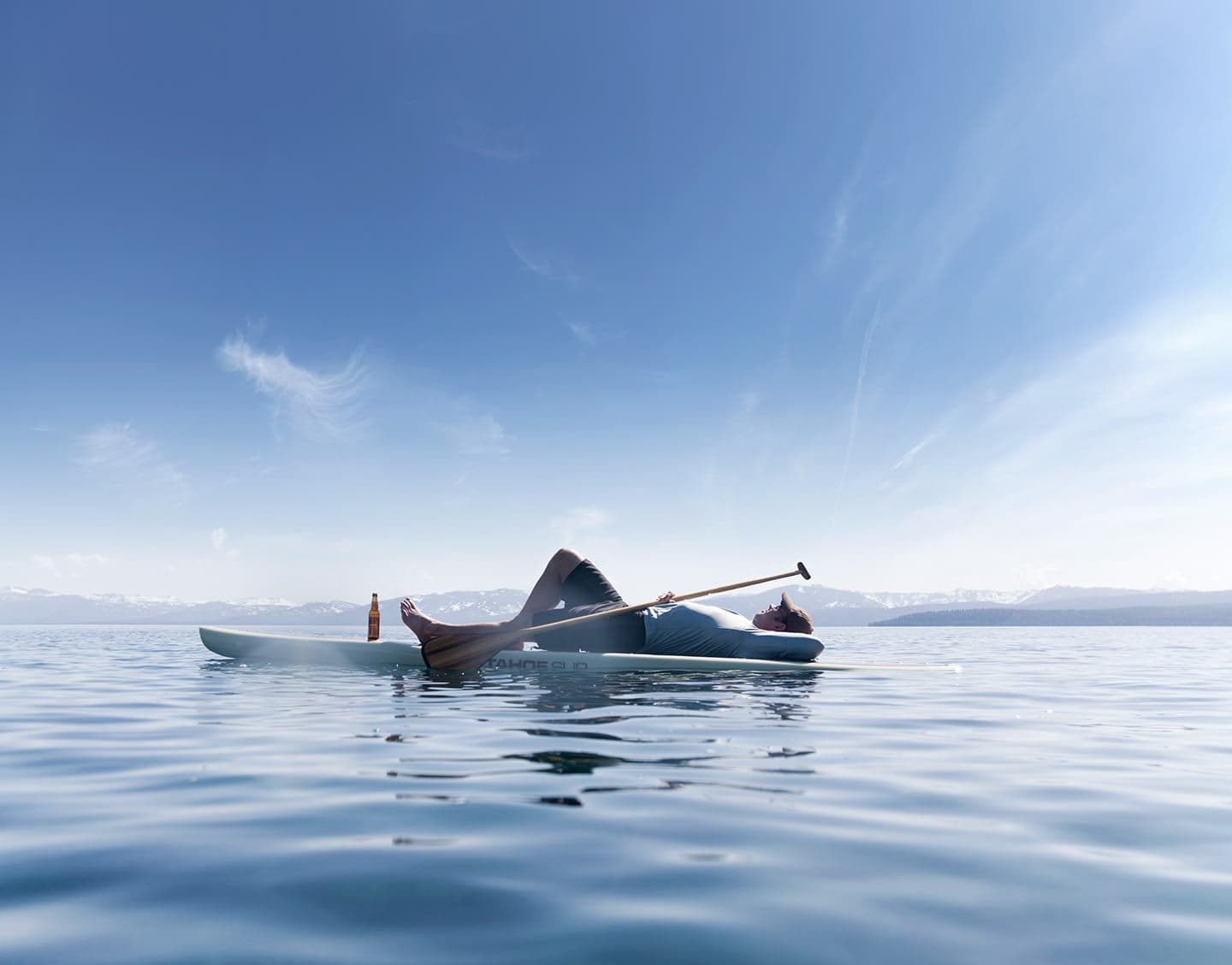 Man-laying-on-paddle-board-on-Tahoe-lake-with-mountains-Rod-McLean_G